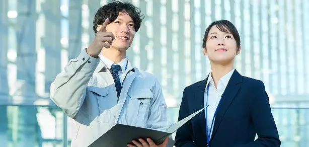 作業服の男性社員と女性社員が建物を指差しながら確認している様子