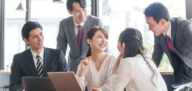 スーツ姿の5名の社員がPCを囲んで笑顔でミーティングしている様子